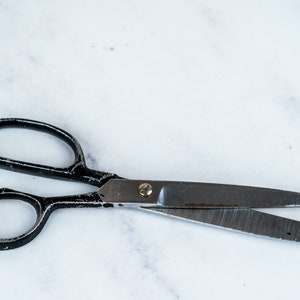 Puede incluir: Tijeras con mangos negros y cuchillas plateadas sobre una superficie de mármol blanco.