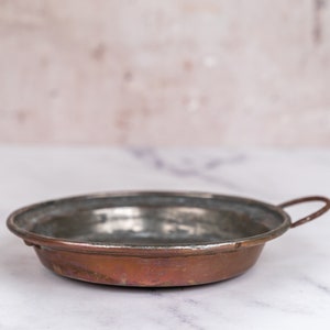 May include: A small, round copper bowl with a single handle. The bowl is slightly tarnished and has a rustic appearance.