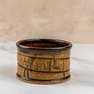 May include: A small, brown ceramic bowl with a dark brown rim and a textured, patterned design. The bowl is sitting on a white marble surface.