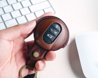 May include: A brown leather key fob with a black key inside. The key has a Mini Cooper logo and a car icon. The fob has a gold ring attached to it.