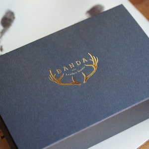 Puede incluir: Una caja gris oscura con un logotipo en relieve dorado que dice "Banda Leather Goods" y presenta un dise&ntilde;o de asta de ciervo dorada estilizada.