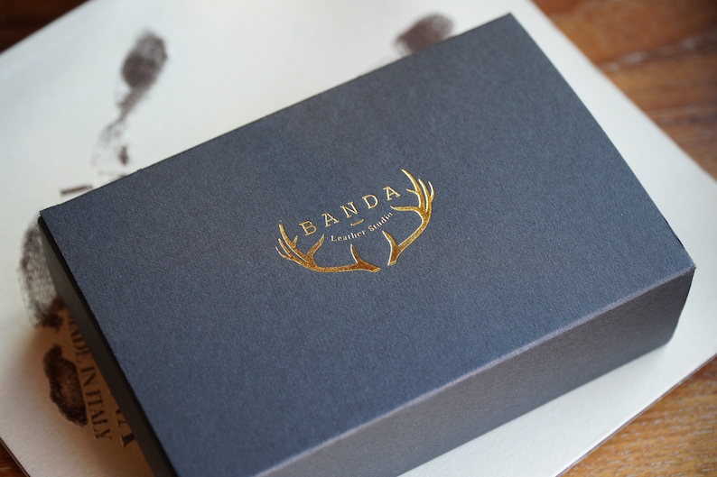 Puede incluir: Una caja gris oscura con un logotipo en relieve dorado que dice "Banda Leather Goods" y presenta un dise&ntilde;o de asta de ciervo dorada estilizada.
