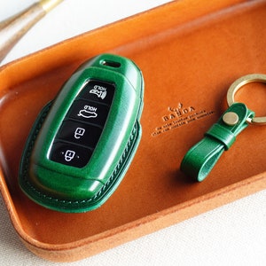 May include: A green leather key fob with four buttons labeled "HOLD" and a silver key ring. The key fob is sitting on a brown leather tray with the text "BANDA" printed on it.