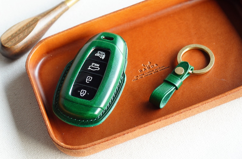 May include: A green leather key fob with four buttons labeled "HOLD" and a silver key ring. The key fob is sitting on a brown leather tray with the text "BANDA" printed on it.