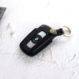 Puede incluir: Un llavero de cuero negro con un logotipo BMW plateado y un anillo de llave dorado. El llavero est&aacute; sobre una superficie blanca.