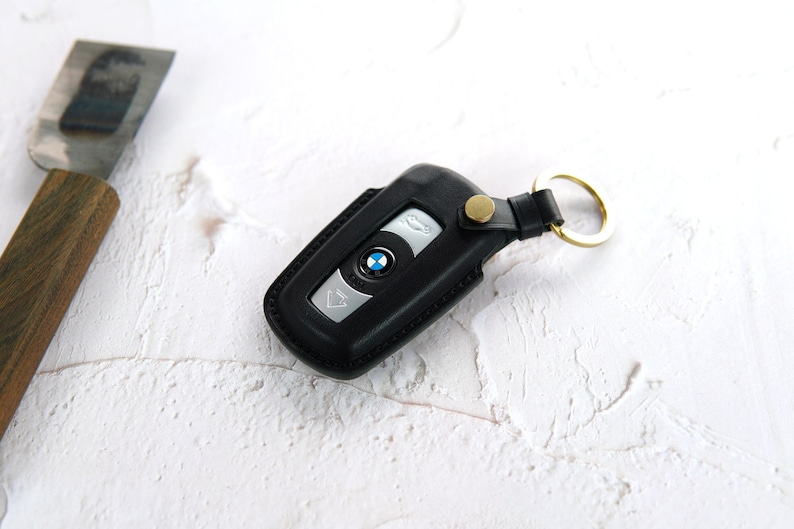 Puede incluir: Un llavero de cuero negro con un logotipo BMW plateado y un anillo de llave dorado. El llavero est&aacute; sobre una superficie blanca.