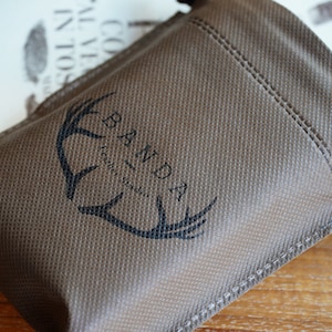 Puede incluir: Una bolsa de tela marr&oacute;n con cierre de cord&oacute;n. La bolsa tiene un logotipo negro que dice "Banda - craft studio" con un dise&ntilde;o de asta de ciervo estilizado.