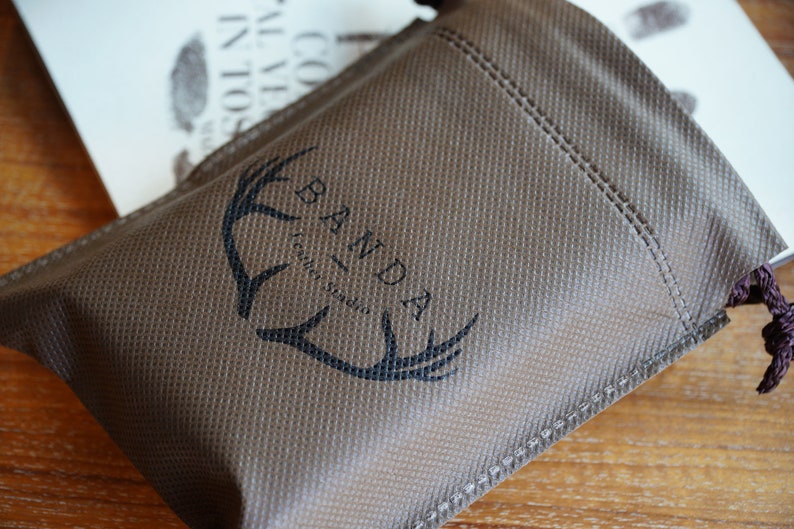 Puede incluir: Una bolsa de tela marr&oacute;n con cierre de cord&oacute;n. La bolsa tiene un logotipo negro que dice "Banda - craft studio" con un dise&ntilde;o de asta de ciervo estilizado.