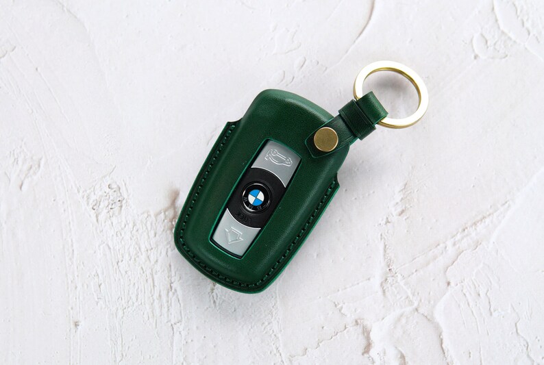 Puede incluir: Un llavero de cuero verde con un anillo de llave dorado y un logotipo BMW plateado.