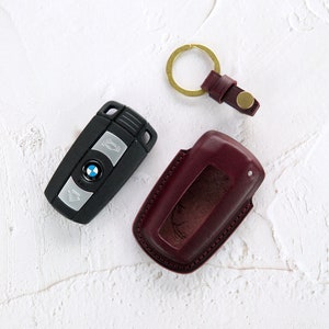 Puede incluir: Una llave de coche negra con un logotipo BMW plateado y un llavero de cuero burdeos con un anillo de llave dorado.