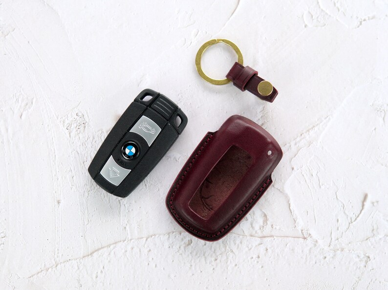 Puede incluir: Una llave de coche negra con un logotipo BMW plateado y un llavero de cuero burdeos con un anillo de llave dorado.