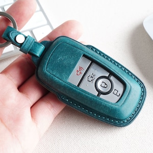Op de afbeelding: Een teal leren sleutelhanger met stiksels rond de randen. De sleutelhanger heeft een zilveren sleutel met knoppen voor vergrendelen, ontgrendelen en paniek. De sleutelhanger heeft een klein rood lampje bovenaan.