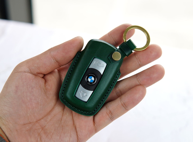 Puede incluir: Un llavero de cuero verde con una llave plateada y un logotipo de BMW. El llavero tiene un anillo de metal dorado.