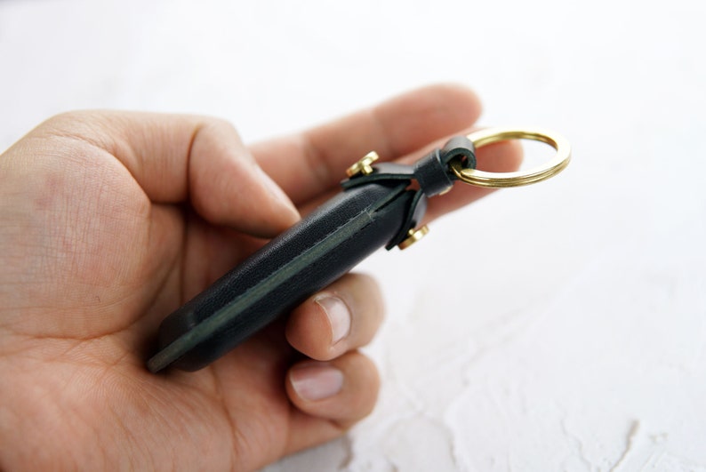 Puede incluir: Un llavero de cuero verde oscuro con un anillo de metal dorado. El llavero tiene una costura cosida en el centro y se sostiene en una mano.