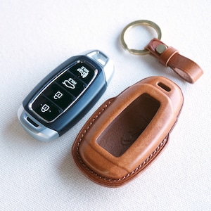 May include: A brown leather key fob with a silver key inside. The key fob has a key ring attached to it.