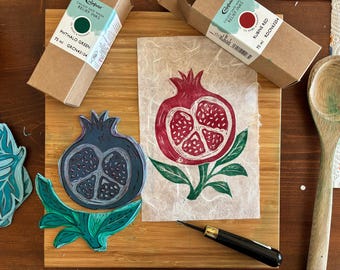 May include: A wooden block print of a pomegranate in red and green ink, with a blue pomegranate print and a green leaf print. Two boxes of Caligo Safe Wash Relief Inks are visible, one labeled "PHTHALO GREEN" and the other "RUBINE RED". A wooden spoon and carving tool are also present.