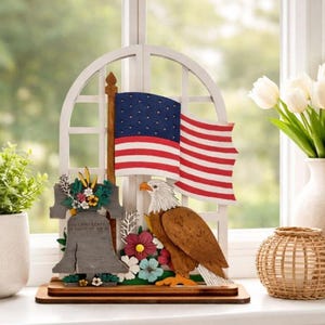 May include: A patriotic wooden display featuring an American flag, a bald eagle, and the Liberty Bell. The flag has red and white stripes with blue stars. The eagle is brown and white, and the bell is gray with floral accents. The display sits on a wooden base.