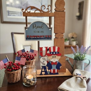 May include: Wooden patriotic sign with the text "Proud to be an American." The sign is accompanied by small American flags, berry-filled baskets, a candle in a jar, and a wooden Uncle Sam figure. A decorative napkin ring with a star is also present.