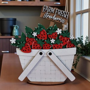May include: A white wooden basket filled with red strawberries, green leaves, and white flowers. A wooden sign reads "FARM FRESH strawberries". The basket has a handle and sits on a wooden base, perfect for kitchen decor.