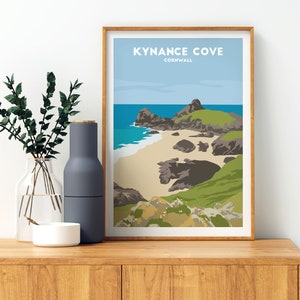 May include: A framed print of a colourful illustration of Kynance Cove in Cornwall, England. The print features a beach with a rocky cliff and blue water.