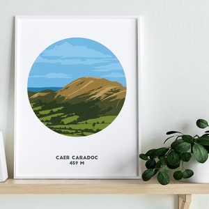 May include: A white framed print with a circular illustration of a green and brown mountain range under a blue sky with white clouds. The text "CAER CARADOC 459 M" is printed below the illustration.