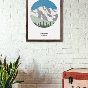 May include: A framed print with a minimalist illustration of a mountain peak with snow and a forest of trees in a circle. The text below the image reads "Jungfrau 4,158 M".