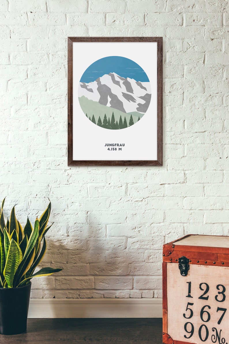May include: A framed print with a minimalist illustration of a mountain peak with snow and a forest of trees in a circle. The text below the image reads "Jungfrau 4,158 M".