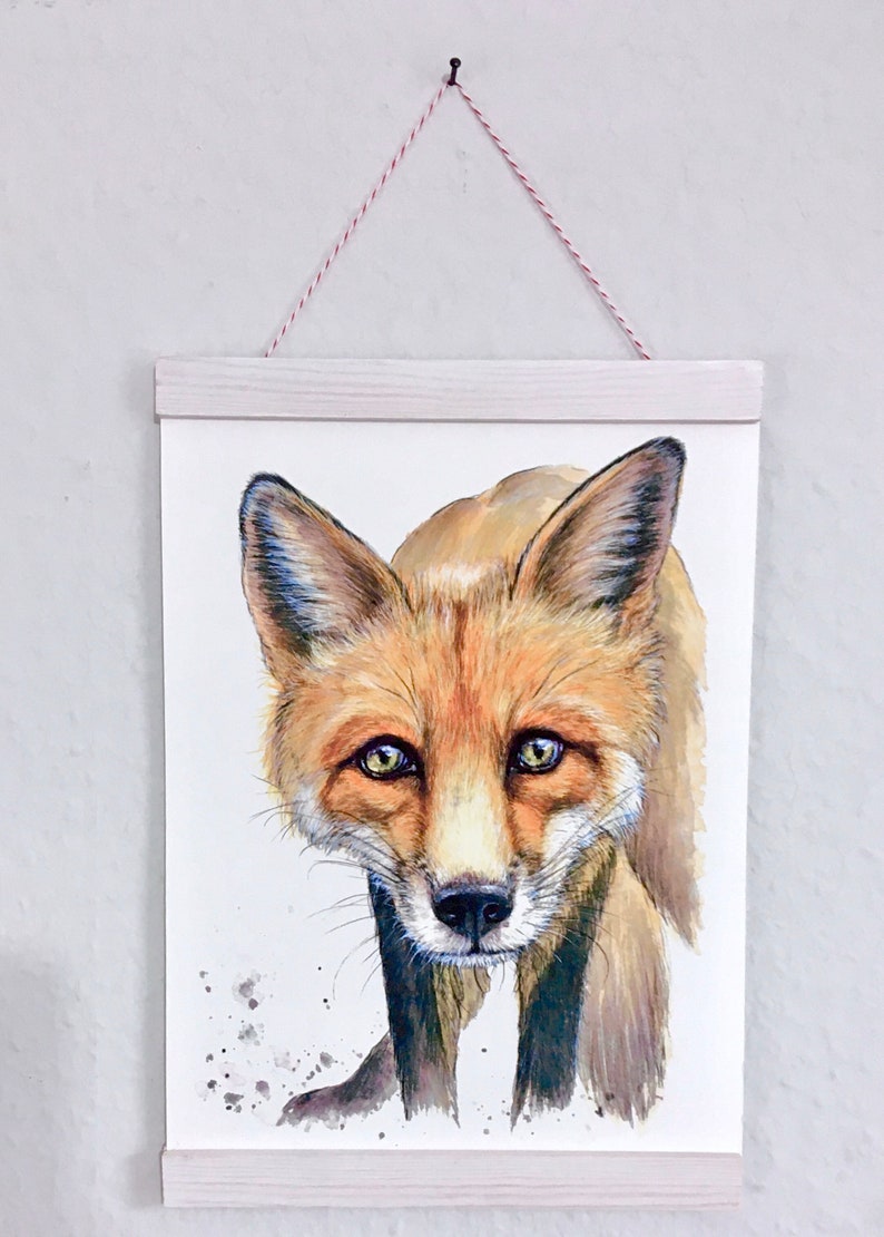 May include: Watercolor painting of a fox with orange and brown fur, green eyes, and a black nose. The artwork is displayed on white paper, held by a light wood frame and suspended by a red and white string.