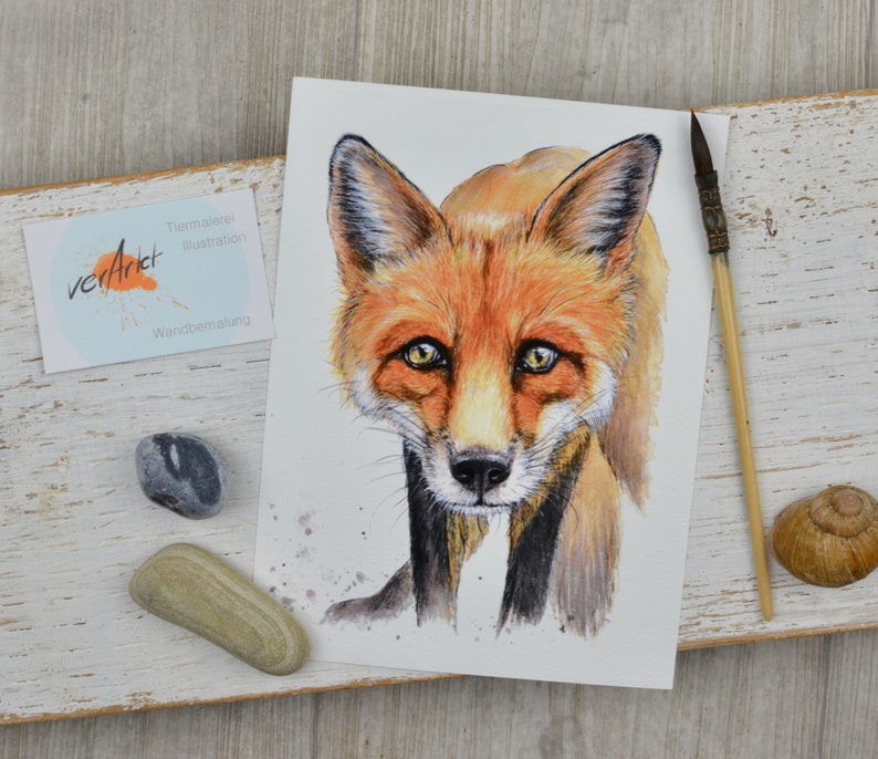 May include: Watercolor painting of a fox with orange and brown fur, yellow eyes, and a black nose. The artwork is on white paper, with the artist's signature and the words "Tier Malerei Illustration Wandbemalung" visible. A paintbrush and a snail shell are next to the artwork.