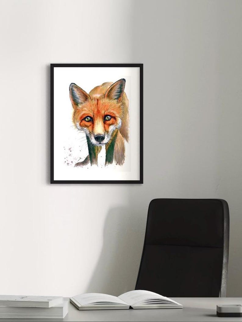 May include: Framed watercolor painting of a fox. The fox has orange fur, a white muzzle, and green eyes. The artwork is in a black frame and is displayed on a white wall. The painting has a realistic style.