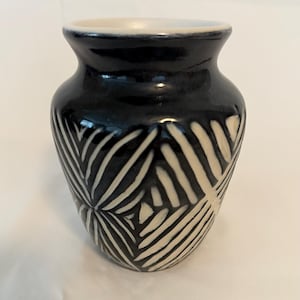 May include: A small, black ceramic vase with a cream-colored rim and a white, geometric leaf pattern. The vase has a rounded body and a narrow neck, suitable for displaying small flowers or decorative items.