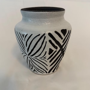 Può includere: Un piccolo vaso in ceramica bianca con bordo nero e motivo geometrico nero. Il vaso ha un corpo arrotondato e un'apertura leggermente svasata. Il motivo nero è composto da linee e forme che creano un design decorativo.