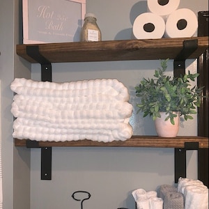 May include: Two wooden shelves with black metal brackets. The top shelf has a white sign that says "Hot 20c Bath Soap & Towels Extra". There is a small clear glass bottle with a white label on the top shelf. The bottom shelf has a stack of white towels, a small potted plant with green leaves, and a stack of white toilet paper rolls.