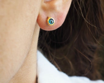 May include: A pair of gold earrings with a blue evil eye charm. The earrings are small and delicate, and the evil eye charm is set in a gold bezel.