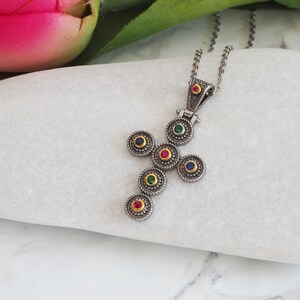 May include: A silver cross pendant necklace with a chain. The cross is decorated with seven small circles, each with a different colored gemstone in the center. The gemstones are red, green, blue, and yellow.