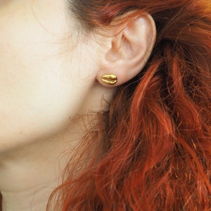 May include: A pair of gold-coloured earrings shaped like a seashell. The earrings are small and delicate, and they are worn by a person with long, red hair.