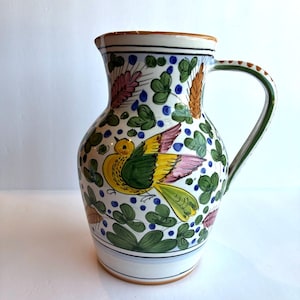 May include: A ceramic pitcher featuring a hand-painted design of a yellow and green bird surrounded by green clover-like leaves, blue dots, and wheat stalks in shades of orange and pink, finished with a brown rim and a green-striped handle.