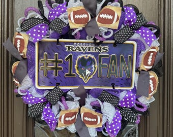 May include: A decorative wreath featuring a Baltimore Ravens license plate with the text "#1 FAN". The wreath includes purple, black, and white mesh, football-shaped ornaments, and ribbons with polka dots.