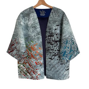 May include: A gray, open-front jacket with ornate calligraphy in various colors. The jacket has a blue lining and is hanging on a wooden hanger. The sleeves are wide and the fabric has a subtle pattern.
