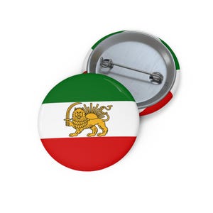 Może przedstawiać: Dwie okrągłe przypinki. Jedna przedstawia irańską flagę z zielonymi, białymi i czerwonymi paskami oraz złotego lwa trzymającego miecz. Druga przypinka pokazuje tył z agrafką.