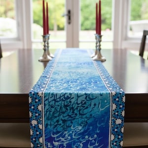May include: A blue table runner with a white floral border and calligraphy design. The runner is placed on a dark brown wooden table. Two silver candle holders with red candles are on the table.