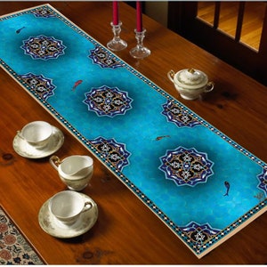 May include: A turquoise table runner with a mosaic design featuring geometric patterns and fish motifs. The runner has a border with a gold and black pattern. The runner is placed on a wooden table, with teacups and a creamer visible.