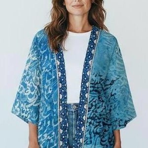 Może przedstawiać: Niebieska kurtka w stylu kimono z białym obramowaniem i wzorem pisma. Kurtka jest noszona na białym topie i niebieskich dżinsach. Rękawy są szerokie, a kurtka jest otwarta z przodu.