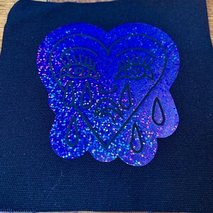 May include: A blue glitter heart-shaped patch with a crying face design. The patch features black outlines and teardrops, set against a black fabric background. The glitter effect adds a sparkling, iridescent quality to the design.