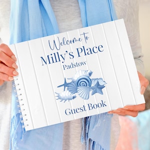 Może przedstawiać: Biała księga gości z niebieskim tekstem i ilustracjami muszli. Na okładce widnieje napis "Welcome to Milly's Place Padstow Guest Book". Książka ma spiralną oprawę. Jasnoniebieski szalik znajduje się w tle.