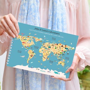 May include: A spiral-bound world map with a teal background, featuring colorful illustrations of landmarks and icons. The text "John & Laura's Adventures" is at the top. The map is held by a person.