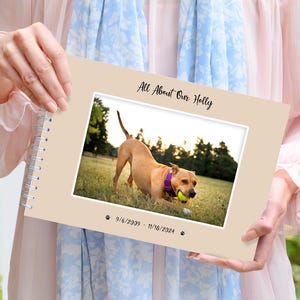 Può includere: Un album fotografico beige con rilegatura a spirale. La copertina presenta una foto di un cane marrone che gioca con una palla da tennis gialla in un campo erboso. Il testo "All About Our Holly" è stampato in corsivo nero sopra la foto. Le date "9/6/2009 - 7/18/2024" sono stampate sotto la foto.