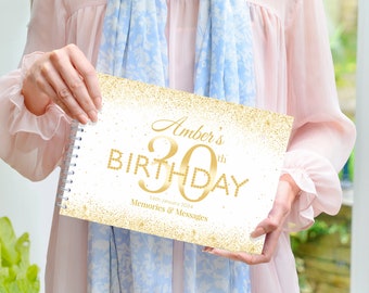 Op de afbeelding: Een wit en gouden verjaardagsgeheugenboek met een gouden glitterachtergrond. De omslag heeft de tekst "Amber's 30th Birthday" met gouden glitteraccenten en de datum "16th January 2024". Het boek heeft een gouden spiraalbinding en de tekst "Memories & Messages" staat onder de datum.