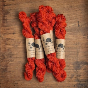 May include: Several skeins of vibrant orange yarn are arranged on a wooden surface. Each skein is wrapped with a paper label featuring a hedgehog illustration and the text "Pinnsvinn Studio 100% laine 25 metres."