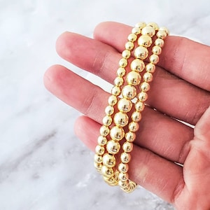 May include: Four gold-toned beaded bracelets stacked on top of each other. The bracelets are made of small, round beads.